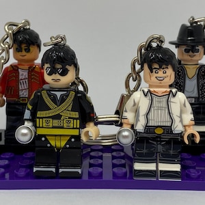 Puede incluir: Cuatro llaveros de minifiguras de plástico sobre una base rectangular de ladrillos morados. Las figuras tienen cabello negro y diversos atuendos, incluyendo una chaqueta roja, un uniforme militar negro y amarillo, una camisa blanca y un traje negro con sombrero.