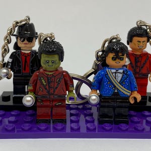 Può includere: Quattro portachiavi con personaggi in plastica e abiti distinti. Uno indossa un completo nero con cappello, uno è un personaggio dalla pelle verde con giacca rossa, uno indossa una giacca blu con paillettes e uno è vestito di rosso con occhi gialli.