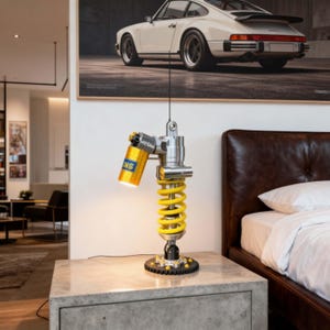 Puede incluir: Una lámpara de mesa construida con un amortiguador de metal plateado que presenta un resorte helicoidal amarillo brillante y un depósito cilíndrico amarillo con el texto OHLINS impreso. La base es un engranaje circular negro con pernos plateados.