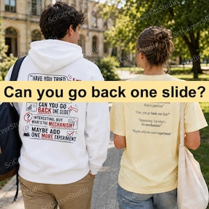 May include: A white hooded sweatshirt and a pale yellow t-shirt, both featuring black and red text prints. The white hoodie displays a list of phrases including "Have you tried," "Can you go back one slide," and "Maybe add one more experiment."