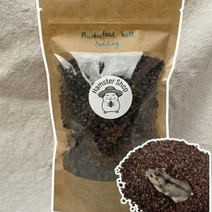 Può includere: Un sacchetto di carta marrone contenente lettiera di pula di grano saraceno marrone scuro. L'etichetta riporta "Buckwheat hull bedding" e un adesivo circolare "Hamster Shop" con l'illustrazione di un criceto. Un piccolo criceto è sulla lettiera.