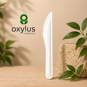 Op de afbeelding: Een wit wegwerpmes met een gekartelde rand staat verticaal. Links is het groene logo van oxylus ecogreen LLC zichtbaar. Het mes is geplaatst naast een gestructureerd beige stenen blok en groene bladeren.