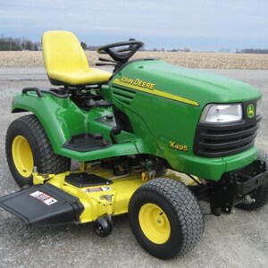 Könnte beinhalten: Ein grüner und gelber John Deere X495 Rasentraktor mit gelbem Sitz, schwarzem Lenkrad und schwarzem Seitenauswurf. Die vordere Motorhaube zeigt das John Deere Logo und die Modellnummer X495 in gelber Schrift.
