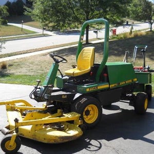 May include: A John Deere F1145 front-mount mower featuring a green chassis, a yellow seat, and a yellow front-mounted cutting deck. The machine includes a roll-over protection structure, black tires with yellow rims, and a rear-mounted utility trailer.