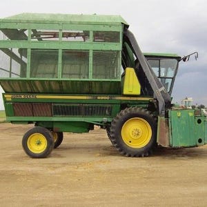 Puede incluir: Una cosechadora de algodón John Deere 9950 de color verde con una gran cesta de almacenamiento de malla elevada, una cabina de conductor con ventanas de cristal y cuatro ruedas grandes con llantas amarillas y un conducto de entrada negro.