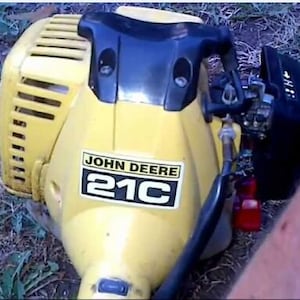 May include: A yellow John Deere 21C string trimmer engine housing featuring a black plastic top cover, a vented side casing, and a black air filter assembly. The unit displays the John Deere brand name and 21C model text on a white label.