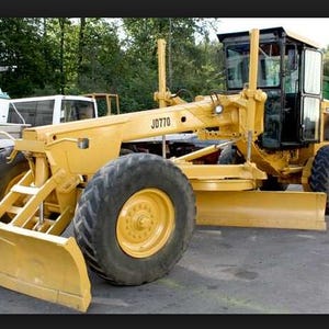 Könnte beinhalten: Ein gelber Motorgrader mit einem großen Frontschild, einem mittig angebrachten Schar und robusten Reifen. Die Baumaschine verfügt über eine schwarze, geschlossene Fahrerkabine und die Aufschrift JD770 ist deutlich am Hauptrahmen zu sehen.