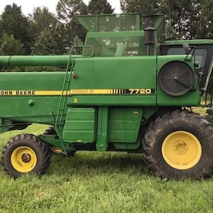 Op de afbeelding: Een groene John Deere 7720 Titan II maaidorser met een grote graantank, een zijladder, een cabine met een zwarte ronde luchtinlaat en gele wielen met zwarte banden. De merknaam en het modelnummer staan op de zijkant van de machine vermeld.