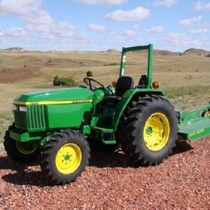 May include: A green John Deere tractor with yellow wheels and a black seat, featuring a roll-over protection structure and a rear-mounted mower attachment. The tractor has a front grille, headlights, and a steering wheel, positioned on a gravel surface.
