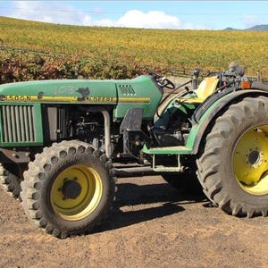 Könnte beinhalten: Ein grüner John Deere 5500N Traktor mit gelben Rädern und einem gelben Sitz. Der Traktor verfügt über einen Frontgrill, große Reifen mit Profil und die Zahl 103, die in Grün auf der Seite der Motorhaube steht. Er steht auf braunem Boden.