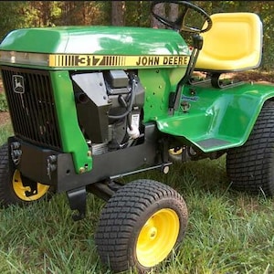 May include: A green and yellow John Deere 317 garden tractor featuring a black front grille, a black steering wheel, and a yellow seat. The tractor has four large, treaded black tyres with yellow rims and is positioned on a patch of green grass.