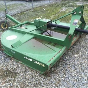 Puede incluir: Una segadora rotativa agrícola John Deere 709 de color verde, con un eje de toma de fuerza negro, un marco de metal y una rueda trasera giratoria. El equipo muestra la marca John Deere y el número de modelo 709 en texto de color blanco.
