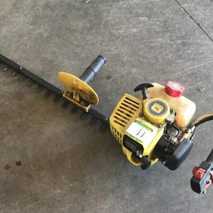 Puede incluir: Un cortasetos de gasolina amarillo y negro con una larga cuchilla de corte de metal, carcasa del motor amarilla con el logotipo de John Deere, depósito de combustible translúcido con tapa roja y mango negro con controles rojos.