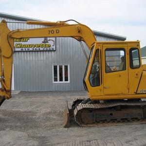 Könnte beinhalten: Ein gelber John Deere 70 D Bagger mit Kettenfahrwerk, hydraulischem Auslegerarm und einer Metallschaufel mit Zähnen. Die Fahrerkabine verfügt über Glasfenster und eine Tür, wobei die Modellnummer 70 D auf dem Auslegerarm aufgedruckt ist.