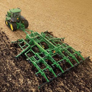 Peut inclure: Un tracteur vert à chenilles en caoutchouc noir tirant un grand cultivateur à dents vert dans un champ. La machine agricole laboure activement le sol, retournant la terre sombre tout en traversant une zone de résidus de culture secs et fauves.