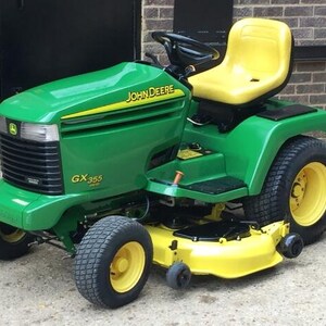 May include: A green and yellow John Deere GX355 lawn tractor featuring a black steering wheel, a yellow seat, a front grille with headlights, and a yellow mower deck. The vehicle has four black tyres with yellow rims and prominent branding.