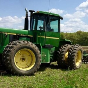 Könnte beinhalten: Ein grüner John Deere 8450 Traktor mit großen schwarzen Reifen und gelben Felgen. Der Traktor verfügt über eine geschlossene Kabine mit getönten Scheiben, ein vertikales schwarzes Auspuffrohr und einen Frontgrill. Er steht auf einem Feld.