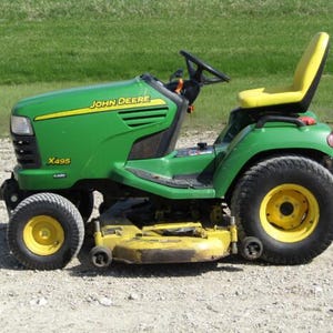 Peut inclure: Un tracteur de pelouse John Deere X495 vert et jaune avec un volant noir, un siège jaune et un plateau de coupe jaune. Le véhicule est équipé de grands pneus noirs avec des jantes jaunes et est placé sur une surface en gravier avec de l'herbe.