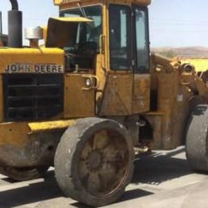 Könnte beinhalten: Ein gelber John Deere Radlader mit einer großen Frontschaufel, einem schwarzen Kühlergrill, einer Fahrerkabine mit Glasfenstern und vier robusten Reifen. Das Baufahrzeug trägt den Markennamen John Deere an der Seite der Motorhaube.