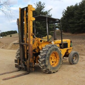 May include: A yellow industrial forklift tractor featuring a tall vertical mast with metal forks, a protective overhead cage, a steering wheel, and large treaded tires. The heavy-duty machinery is painted in a bright yellow finish with black accents.