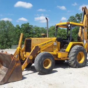 Può includere: Una terna John Deere 710C gialla dotata di benna anteriore, braccio escavatore posteriore, griglia nera e grandi pneumatici tassellati. Il pesante veicolo da costruzione è posizionato su una superficie di ghiaia sotto un cielo azzurro.