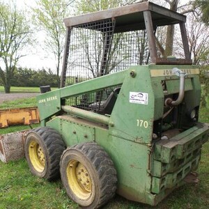 Può includere: Una minipala John Deere 170 di colore verde con una gabbia protettiva in metallo, ruote gialle con pneumatici neri e una benna montata frontalmente. La macchina mostra il marchio John Deere e il numero di modello 170 sui pannelli laterali.