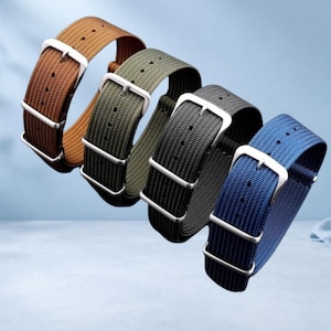 May include: Four nylon watch straps arranged in a diagonal row, featuring brown, olive green, black, and blue colors. Each strap includes a silver-toned metal buckle and two matching metal keepers, with a woven texture and pre-punched adjustment holes.
