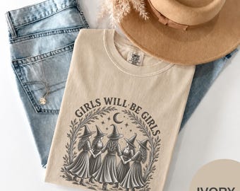 May include: An ivory t-shirt featuring a dark gray graphic of five figures in pointed hats holding hands in a circle under a crescent moon and stars, with the text "GIRLS WILL BE GIRLS" arched above, paired with light blue denim jeans and a tan hat.