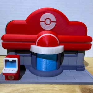 Op de afbeelding: Een 3D-geprint model van een rood, wit en blauw gebouw met een cirkelvormig embleem op het dak. De structuur bevat een centrale blauwe deur, een grijze basis en een kleine rode kiosk met een blauw scherm links van de ingang.