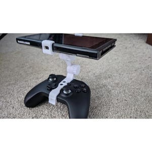 Puede incluir: Un soporte de montaje de plástico blanco unido a un mando de videojuegos negro, con un brazo ajustable que sostiene una consola de juegos portátil negra en posición horizontal sobre el mando para una visualización integrada.