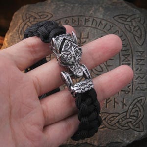 May include: A black paracord bracelet featuring a silver-toned metal clasp shaped like a stylized wolf head with intricate engraved knotwork patterns. The cord is woven in a thick, braided design, connecting to the metallic animal head centerpiece.