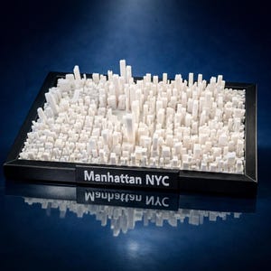May include: A three-dimensional white model of the Manhattan NYC skyline featuring numerous rectangular building structures of varying heights, all contained within a black rectangular frame with the text Manhattan NYC on the front edge.