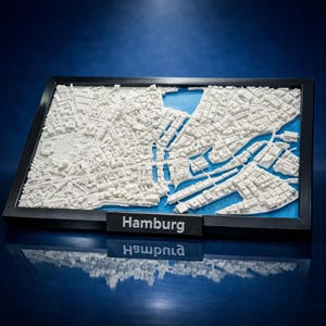 Peut inclure: Une carte en relief 3D encadrée de Hambourg présentant des structures architecturales blanches sur un fond bleu. Le cadre noir comprend une petite plaque rectangulaire au centre en bas avec le texte blanc Hamburg imprimé dessus.