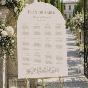 Peut inclure: Un panneau de plan de table de mariage blanc en forme d'arche avec une inscription dorée Plan de Table, Sarah & Clément, et 18/07/2026. Il liste douze tables avec des noms en gris et présente un dessin floral linéaire décoratif en bas.