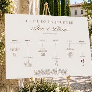 Könnte beinhalten: Ein weißes, rechteckiges Hochzeitsschild auf einer goldenen Staffelei mit dem Text LE FIL DE LA JOURNÉE, Alex & Léana und 12 septembre 2026. Das Schild zeigt einen chronologischen Zeitplan mit Symbolen für Zeremonie und Empfang.