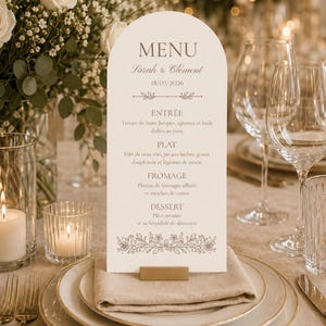 Può includere: Un cartoncino menu bianco ad arco con testo dorato che riporta MENU, Sarah & Clément, 18/07/2026 e un elenco di portate tra cui Entrée, Plat, Fromage e Dessert, con un'illustrazione floreale sul fondo, sostenuto da un piccolo supporto dorato.