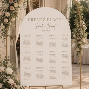 Peut inclure: Un panneau de plan de table de mariage blanc en forme d'arche avec le texte PRENEZ PLACE, Sarah & Clément, et la date 18/07/2026. Il liste douze tables avec des noms d'invités, sur un support en métal doré orné de roses blanches et feuillage.