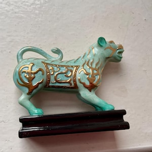 May include: A turquoise ceramic tiger figurine featuring gold-coloured decorative patterns on its body, including a rectangular saddle-like design on its back. The tiger stands on a rectangular, dark brown wooden base with a smooth, polished finish.