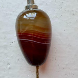 Könnte beinhalten: Ein polierter, tropfenförmiger Achatstein mit horizontalen Bändern in Dunkelbraun, Bernstein und Weiß. Der Stein ist mit einer silberfarbenen Metallkappe mit einem kleinen roten Akzent an der Spitze versehen und an einer dünnen, gedrehten goldfarbenen Kordel befestigt.