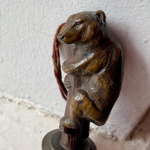 May include: A bronze-toned metal figurine of a bear climbing a tree trunk, mounted on a circular base. The bear is depicted in a realistic pose with textured fur details. A frayed, reddish-brown cord is attached to the side of the bear figure.