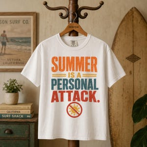 May include: A white short-sleeve t-shirt featuring the distressed orange, teal, and red text "SUMMER IS A PERSONAL ATTACK." above a red circle with a diagonal line through a yellow sun icon. The shirt is displayed on a wooden hanger.