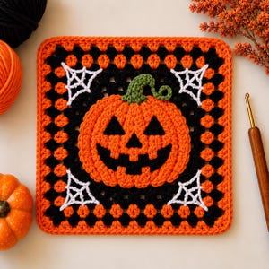 May include: A square crochet granny square featuring a central orange jack-o'-lantern with a green stem, black eyes, and a black mouth. The square is bordered by alternating black and orange yarn, with white spiderweb motifs in each corner.