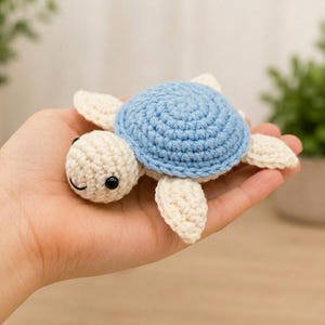 May include: A small crochet turtle toy featuring a light blue shell and cream-colored head, flippers, and body. The turtle has two black plastic bead eyes and a simple stitched black mouth, crafted with a textured yarn stitch pattern throughout.