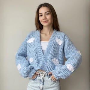 May include: A light blue crochet cardigan featuring a V-neckline, long sleeves with ribbed cuffs, and a pattern of small, white, textured clouds scattered across the front and sleeves. The garment has a cropped length and a thick, chunky knit texture.