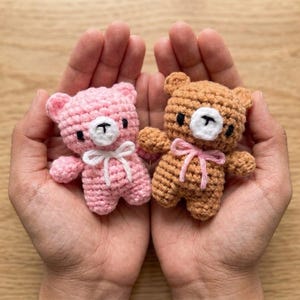 May include: Two small crocheted teddy bears, one light pink and one light brown, each featuring a white muzzle, black embroidered eyes, a black nose, and a small yarn bow tied around the neck in a contrasting colour to the bear's body.