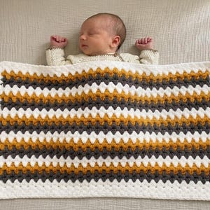 May include: A rectangular crochet baby blanket featuring a repeating striped pattern in mustard yellow, charcoal gray, and white. The blanket is constructed with a textured stitch pattern and finished with a simple white border around the edges.