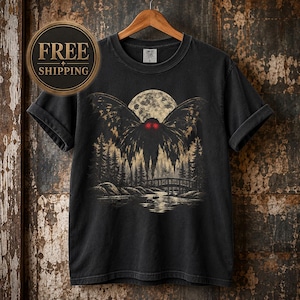 May include: A black short-sleeve t-shirt featuring a graphic print of a winged creature with glowing red eyes hovering above a bridge, a forest, and a river under a large full moon. The shirt has a crew neckline and is displayed on a wooden hanger.