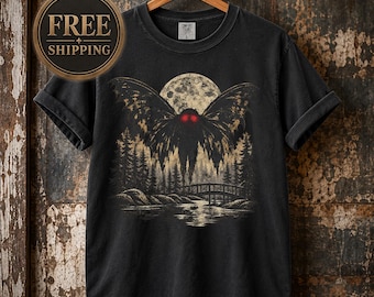 May include: A black short-sleeve t-shirt featuring a graphic print of a winged creature with glowing red eyes hovering above a bridge, a forest, and a river under a large full moon. The shirt has a crew neckline and is displayed on a wooden hanger.
