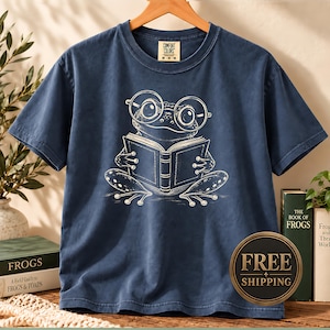 May include: A navy blue short-sleeve t-shirt featuring a white line-art graphic of a frog wearing round glasses and reading an open book. The shirt has a crew neckline and a Comfort Colors brand label printed on the inside of the collar area.