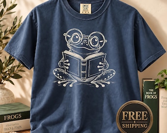 May include: A navy blue short-sleeve t-shirt featuring a white line-art graphic of a frog wearing round glasses and reading an open book. The shirt has a crew neckline and a Comfort Colors brand label printed on the inside of the collar area.